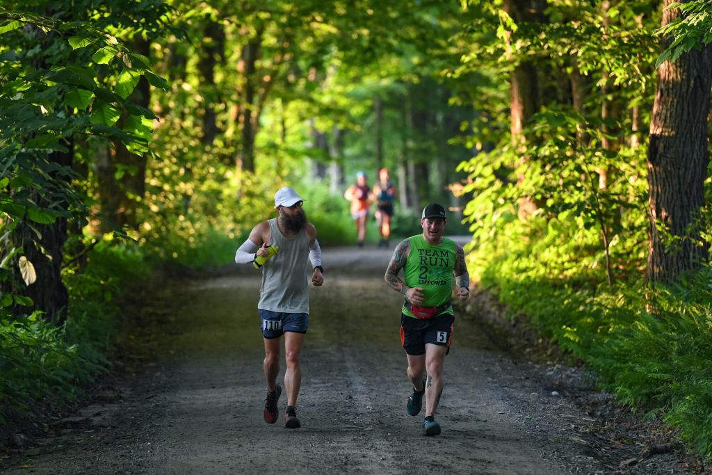 Two runners racing on a dirt road at the Vermont 100. The runner on the looker's right is wearing a Team Run 2 Empower shirt.